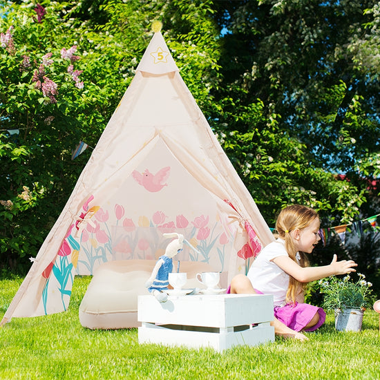 A little girl is sitting next to a pink kid's tent in the grass outdoors.